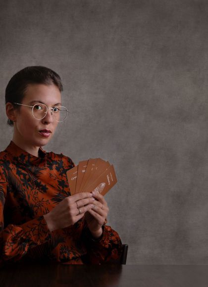 Photo of author Maude White. The author wears glasses and holds cards from the Pleasure Alchemy deck. She has a mysterious look on her face.