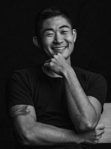 Black and white photo of artist Yoshino from the waist up. The artist is wearing a black shirt, and has one arm crossed in front with the other elbow sitting on that arm and raised up to his chin. He is smiling into the camera.